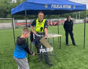 Na zdjęciu stoisko bytomskich mundurowych, przy nim policjantka i policjant, który trzyma policyjną tarczę, kilkuletni chłopiec uderza w nią pałką służbową.