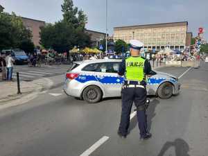 Na zdjęciu policjant drogówki stojący przed radiowozem i obserwujący zgromadzone osoby