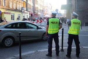 Na zdjęciu dwóch policjantów przy radiowozie służbowym obserwujących zgromadzone osoby