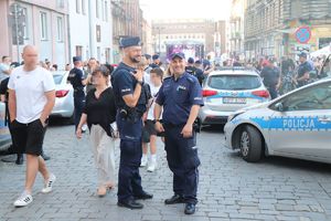 Na zdjęciu dwóch uśmiechniętych policjantów przy radiowozach służbowym obserwujących zgromadzone osoby