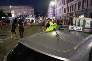 Na zdjęciu dwóch policjantów przy radiowozie służbowym obserwujących zgromadzone osoby