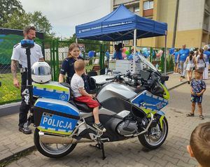 Na zdjęciu policjant, policjantka, motocykl policyjny na którym siedzi kilkuletni chłopiec, obok stoisko i zgromadzone osoby.