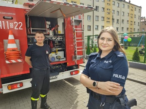 Na zdjęciu policjantka i strażak, za nimi wóz strażacki.