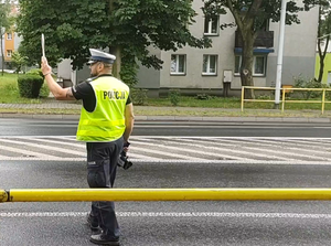 Na zdjęciu widzimy policjanta stojącego na jezdni, ma uniesioną do góry rękę, w której trzyma tarczę do zatrzymywania pojazdów.