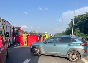 Na zdjęciu samochód, wozy strażackie, policjanci i strażacy na autostradzie, widoczny parawan czerwonego koloru