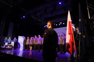 Na zdjęciu stojący na scenie policjanci obok policjant stojący z boku.