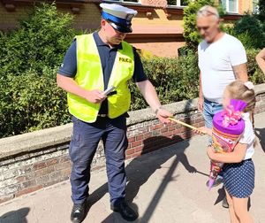 Na zdjęciu policjant bytomskiej drogówki przekazujący żółtą odblaskową opaskę dziewczynce trzymającej w ręku tytę.