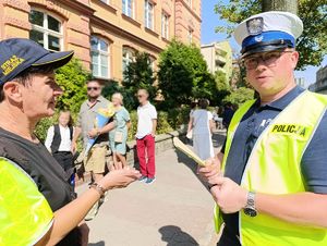 Na zdjęciu policjant bytomskiej drogówki wraz ze strażniczką miejską, przed budynkiem szkoły, dookoła nich przemieszczjące się osoby.