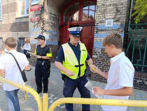 Na zdjęciu policjant bytomskiej drogówki wraz ze strażniczką miejską, przed budynkiem szkoły, dookoła nich przemieszczjące się osoby.