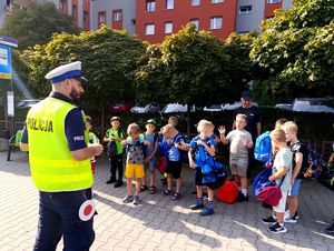 Na zdjęciu policjant drogówki, przed nim dzieci stojący na chodniku.