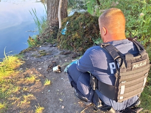 Na zdjęciu policjant przy stawie, przed nim leżące na ziemi granaty, pociski, wiaderko.