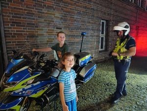 Na zdjęciu policjant, siedzący na motocyklu służbowym chłopiec, obok stojąca dziewczynka.