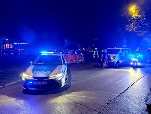 Na zdjęciu radiowóz i motocykl policyjny z włączonymi światłami niebieskiego koloru, ambulans i zgromadzone osoby.