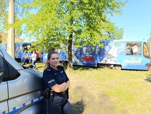 Na zdjęciu policjantka stojąca przy radiowozie służbowym, za nią tramwaj niebieskiegokoloru.