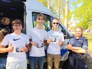 Na zdjęciu trzy osoby trzymające w jednej dłoni ulotki, druga dłoń przedstawia uniesione w górę kciuki, przy nich policjantka.