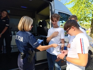 Na zdjęciu policjantka rozdająca ulotki, obok w radiowozie policyjnym kolejna policjantka.