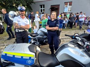 Na zdjęciu policjant drogówki obok policjantka, stoją przed motocyklem służbowym dookoła zgromadzona osoby.