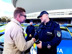 Na zdjęciu umundurowany policjant stojący przy mężczyźnie w jasnym płaszczu, koloru kremowego, trzymają dłonie w uścisku, w trakcie rozmowy.