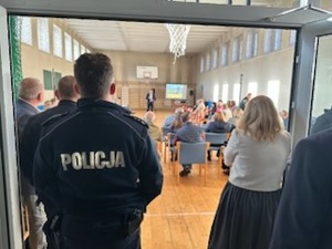 Na zdjęciu policjant stojący tyłem i zgromadzone na sali osoby.