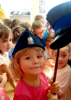 Na zdjęciu dziewczynka w policyjnej czapce, trzyma w ręku policyjną pałkę na której jest kolejna policyjna czapka.