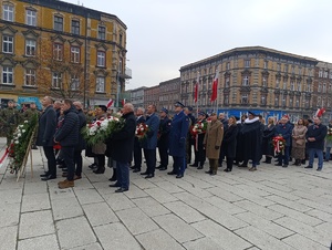 Zgromadzone osoby trzymające w dłoniach biało czerwone kwiaty.