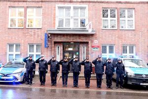 Na zdjęciu policjanci bytomskiej komendy ustawieni w szeregu przed budynkiem komendy, obok nich radiowozy z włączonymi sygnałami świetlnymi.