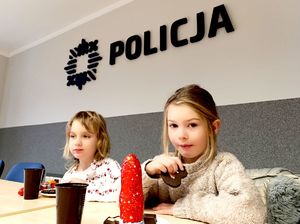 Na zdjęciu dwie dziewczynki siedzące przy stole, jedzące ciastka, za nimi na ścianie napis Policja