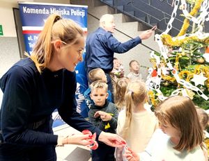 Na zdjęciu dzieci i policjantka rozdająca białoczerwone lizaki choinkowe, za nią policjant ozdabiający z dziećmi choinkę