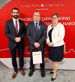 na zdjęciu dwóch mężczyzn i kobieta, stojący w środku mężczyzna ma załażoną odznakę, trzyma w dłoniach torębkę na której widnieje napis bohaterowie