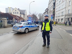 na zdjęciu policjant na jezdni, za nim radiowóz i poruszający się orszak