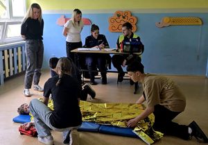na zdjęciu chłopiec leżący na materacu przy nim trójka dzieci udzielająca mu pomocy przykrywając kocem termicznym