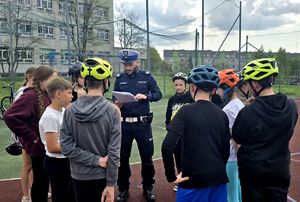na zdjęciu znajdujące się na boisku dzieci, część w założonych na głowach kaskach, przy nich policjant ruchu drogowego