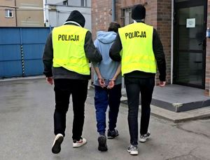 na zdjęciu dwóch policjantów w żółtych kamizelkach, prowadzących zatrzymanego mężczyznę