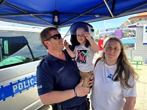 na zdjęciu policjant trzymający małego chłopca na rękach, który ma na głowie kask policyjny, obok nich kobieta