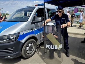 na zdjęciu obok radiowozu służbowego mały chłopiec ubrany w kask, kamizelkę policyjną, trzymający pałkę tarczę policyjną, przy nim policjantka przytrzymuje mu tarczę i pałkę służbową