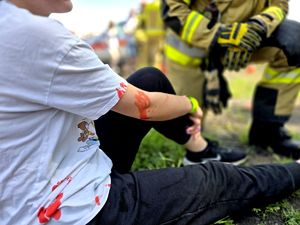 na zdjęciu kobieta siedząca na ziemi z pomalowaną na czerwono ręką i bluzką, przy niej strażak