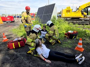 na zdjęciu strażacy i leżące przy nich torby koloru czerwonego, jeden trzyma zabandażowaną głowę kobiety siedzącej na ziemi, drugi pochyla się nad kolejną siedzącą na ziemi kobietą, obok nich kolejny strażak.