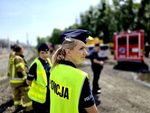 na zdjęciu policjantka, za nią rozmyte tło na którym widoczni są kolejni mundurowi i wóz strażacki