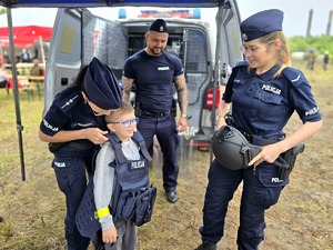 na zdjęciu policjantka trzyma kamizelkę policyjną w którą ubrany jest kilkuletni chłopiec, obok kolejna policjantka, za nimi policjant i radiowóz.