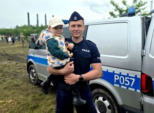 na zdjęciu policjant trzymający małe dziecko na rękach, za nimi radiowóz..
