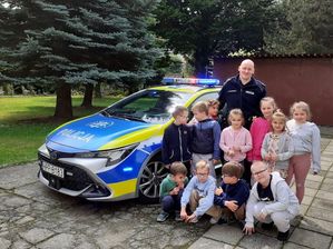 na zdjęciu policjant stojący przy dzieciach obok radiowozu służbowego