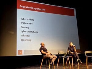 na scenie siedząca na krześle policjantka trzyma przy ustach mikrofon, obok mężczyzna, za mini wyświetlone napisy zagrożenia społeczne, cyberstalking, trollowanie, flaming, cyberprostytucja, seksting, grooming