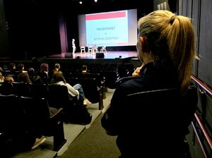 policjantka odwrócona plecami, obok siedzące na sali osoby, na scenie dwie osoby, za nimi napis świadomość równa się bezpieczeństwo