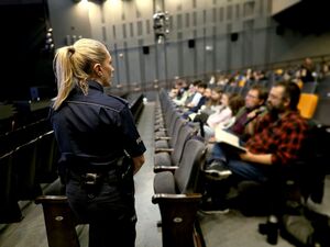 policjantka odwrócona plecami, obok siedzące w sali osoby, mężczyzna przemawia do mikrofonu, który trzyma w dłoni