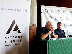 na zdjęciu zastępca komendanta przemawia do mikrofonu, obok mundurowy, siedzą przy stole, obok nich rollup z napisem aktywne śląskie portal obywatelski