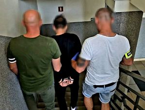 na zdjęciu dwóch mężczyzn trzyma za ramiona mężczyznę w środku, w kajdankach, wszyscy odwróceni tyłem