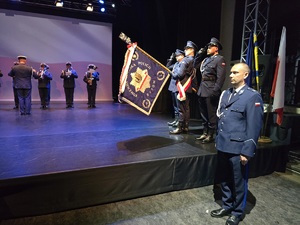 Na zdjęciu widzimy trzech salutujących policjantów, mundurowy w środku trzyma sztandar, obok policjant prowadzący uroczystość stoi na baczność
