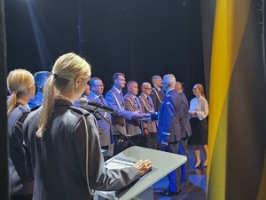 dwie policjantki przy mównicach, obok stojący na scenie mundurowi podczas gratulacji od komendanta wojewódzkiego i miejskiego