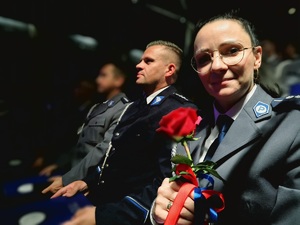 na zdjęciu policjantka z różą w dłoni obok policjant, siedzą na sali