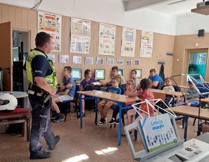 na zdjęciu policjant stojący przed dziećmi siedzącymi w ławkach szkolnych